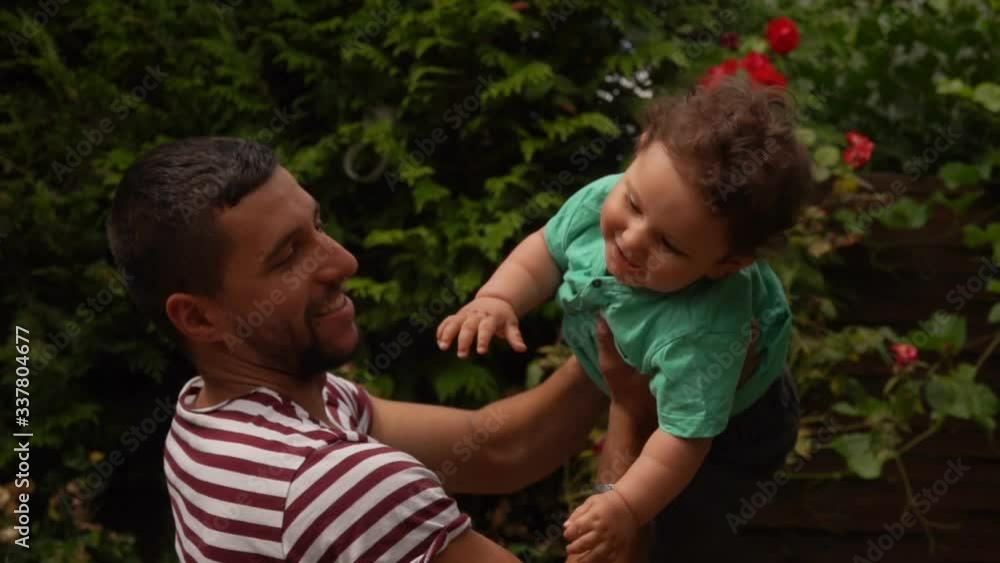 Handsome dad in a striped T-shirt plays and kisses a curly happy baby boy outdoors in the garden Handsome dad in a striped T-shirt plays and kisses a curly happy baby boy outdoors in the garden