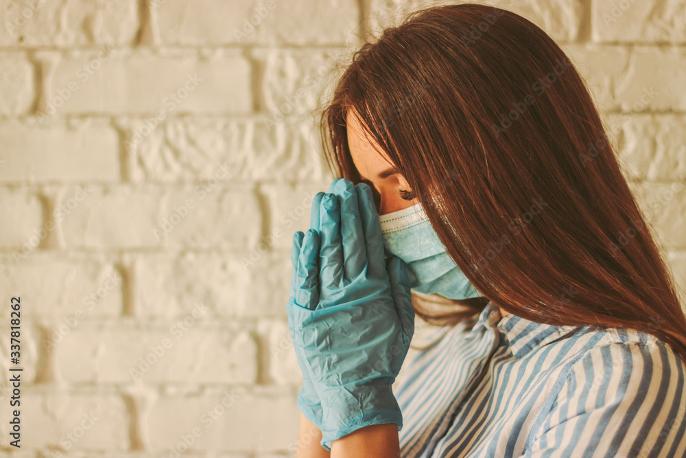 Young faithful woman in protective gloves and medical face mask praying with hope for help at home. Religious girl prayer in protective face mask and medical gloves in church. Coronavirus COVID-19 Young faithful woman in protective gloves and medical face mask praying with hope for help at home. Religious girl prayer in protective face mask and medical gloves in church. Coronavirus COVID-19