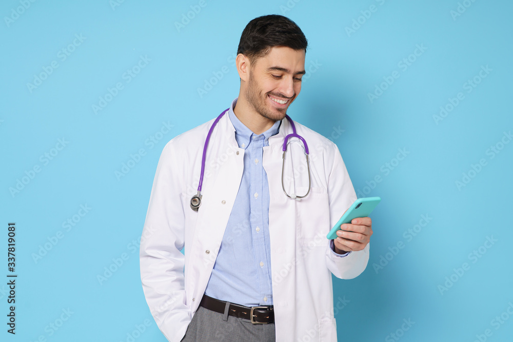 Young handsome doctor in white coat smiling while looking at screen of his phone, using medical app, standing isolated on blue background Young handsome doctor in white coat smiling while looking at screen of his phone, using medical app, standing isolated on blue background