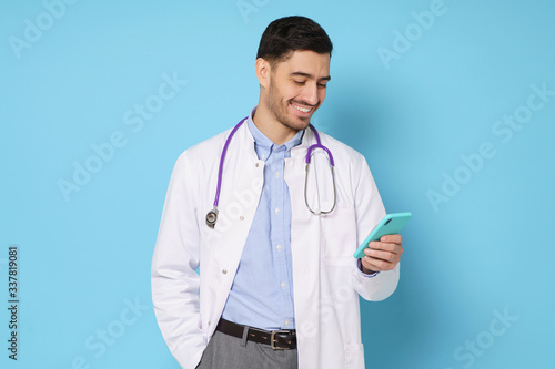 Young handsome doctor in white coat smiling while looking at screen of his phone, using medical app, standing isolated on blue background