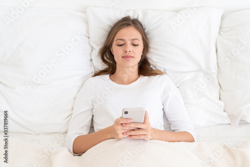 Young woman lying in bed on pillows, wearing white clothes, holding smartphone with both hands and reading posts or messages after awakening in the morning