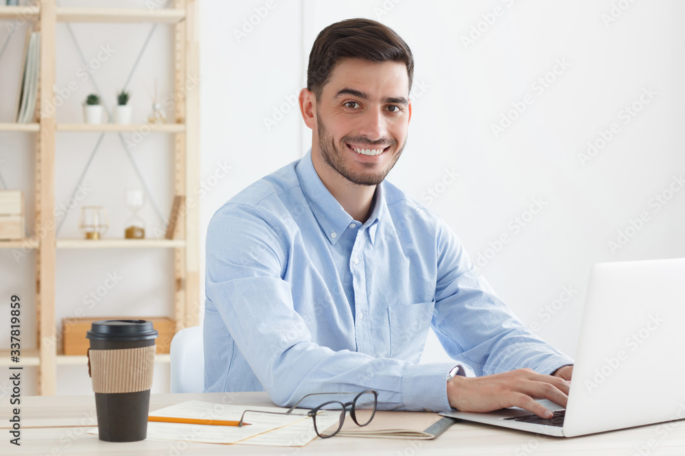Happy smiling male sitting at desk in coworking office, drinking coffee from takeaway cup and doing work on his laptop, looking at camera Happy smiling male sitting at desk in coworking office, drinking coffee from takeaway cup and doing work on his laptop, looking at camera