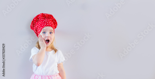 Little blonde smiling girl in red cook's hat staying on neutral grey background and holding her cheek, surpised, shocked. Space for text