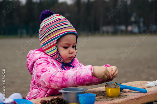 Little girl play with set of children's toy colorful dishes, cones and chestnutson on the bench. Kid toy utensil for girls game. Early spring photo