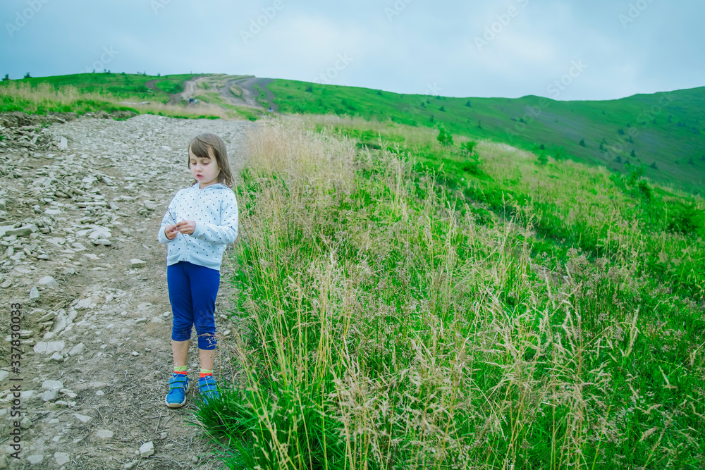 Cute little child girl hiker traveler walks on top of mountain among grass against a backdrop of hills and mountains.