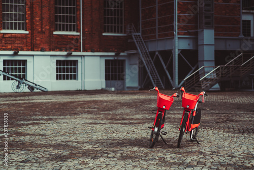 Two vivid red rental bikes parked on a paving stone in front of a brick building; two shared bicycles with metal baskets at the helm standing outdoors on the pavement, with copy space area on the left