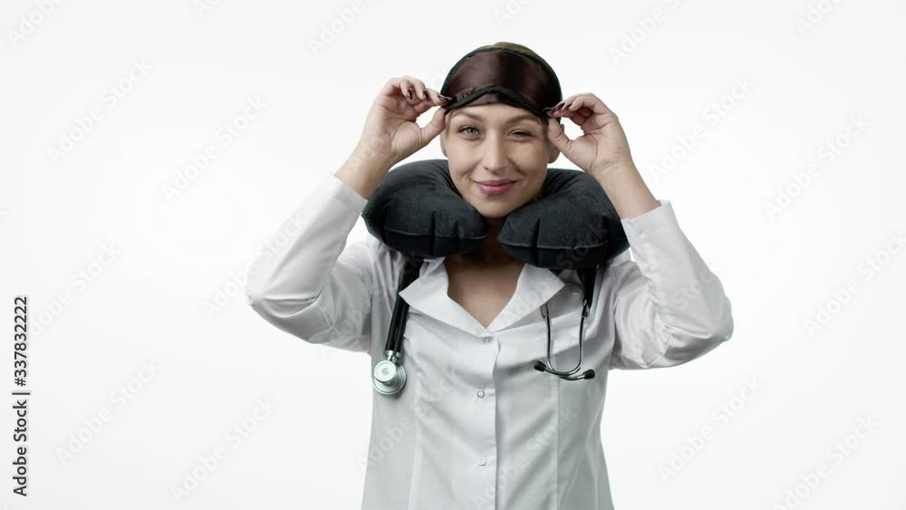 Beautiful sexy female doctor in medical uniform with stethoscope puts on sleeping mask and portable travel neck pillow, playfully winks and fall asleep. Sleeping at work, taking nap, resting concept Beautiful sexy female doctor in medical uniform with stethoscope puts on sleeping mask and portable travel neck pillow, playfully winks and fall asleep. Sleeping at work, taking nap, resting concept