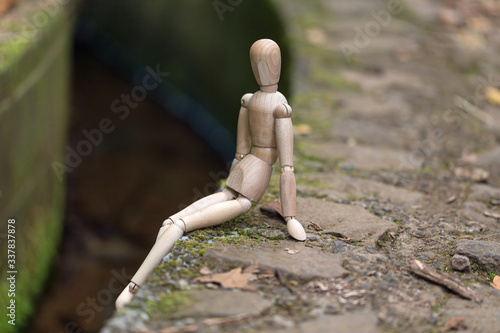 A wooden man sits resting on the edge of a canal in the woods