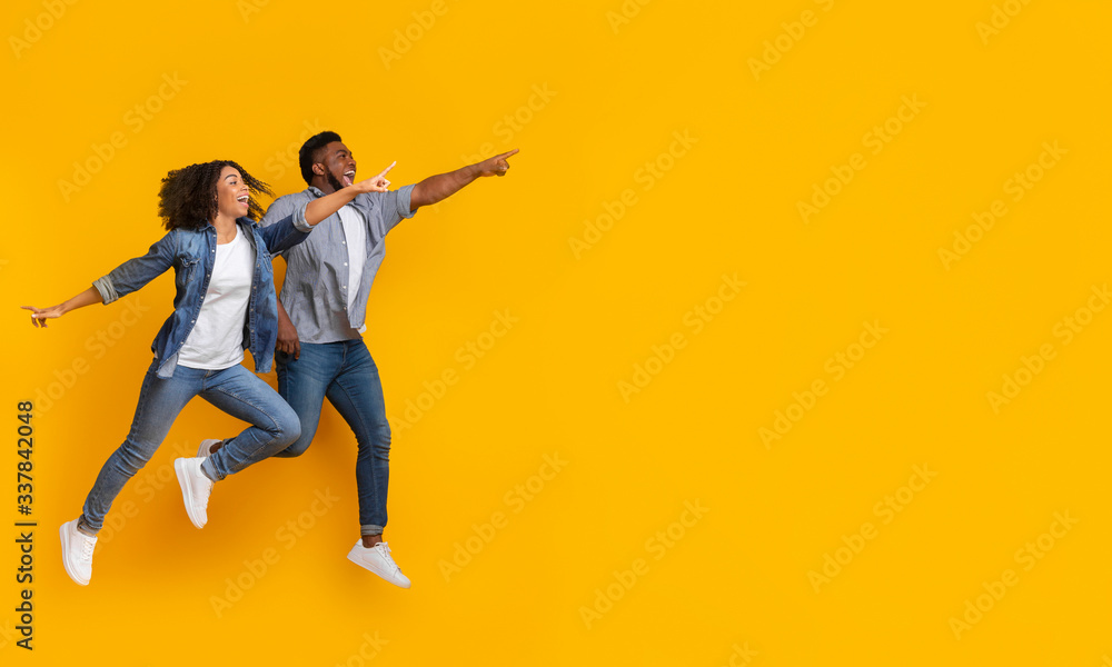 Positive black couple jumping in air and pointing at copy space