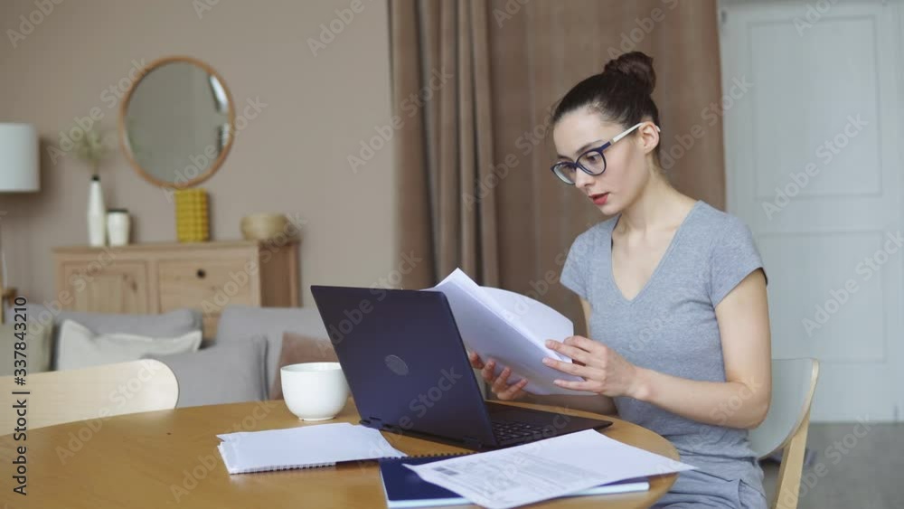 Young woman with glasses reading documents while working on a laptop at home. Distance work or online education. Stay home while quarantined against coronavirus. Young woman with glasses reading documents while working on a laptop at home. Distance work or online education. Stay home while quarantined against coronavirus.