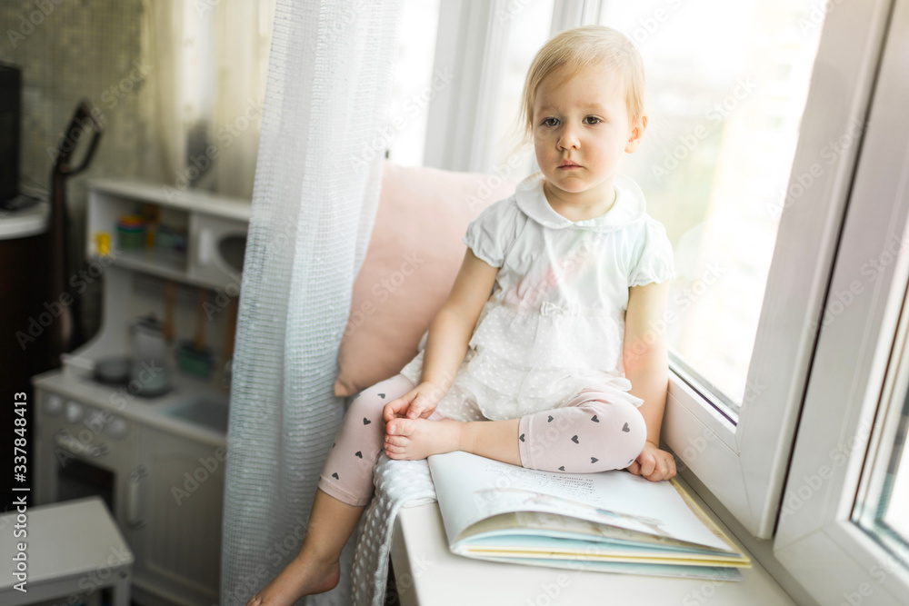 Little girl sitting on the windowsill of the house, self-isolation at home in 2020 Little girl sitting on the windowsill of the house, self-isolation at home in 2020