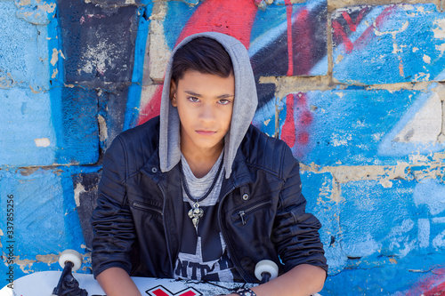 Stylish teenager boy wearing hood shirt with skateboard in city graffiti background looking at you camera sitting photo