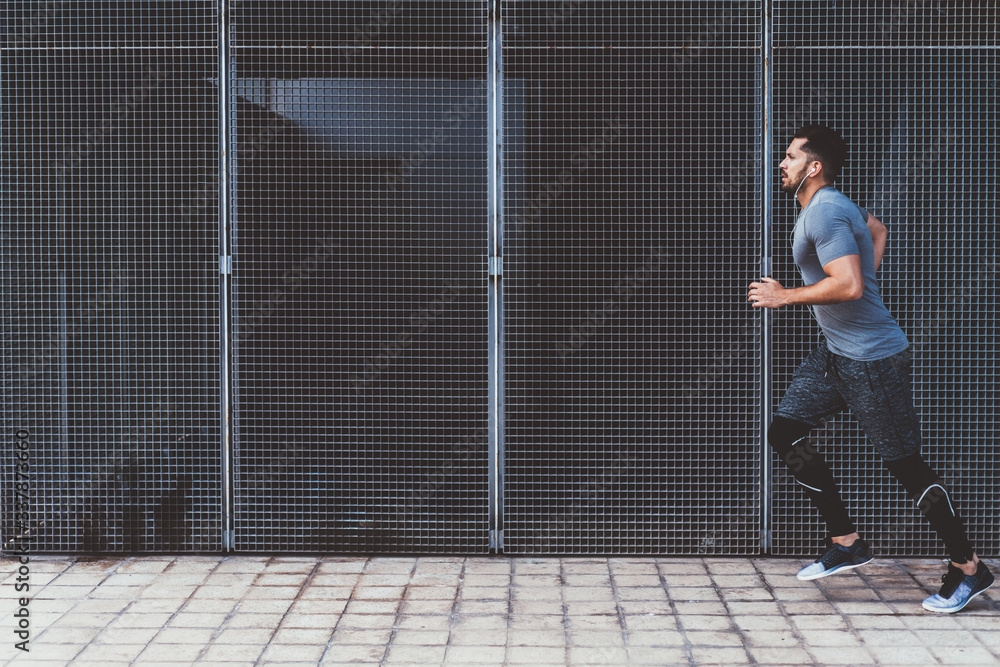 Fototapeta premium Handsome strength male in active wear jogging outdoors on street in earphones listening playlist songs, sportsman having cardio training outdoor keeping healthy lifestyle and perfect body shape