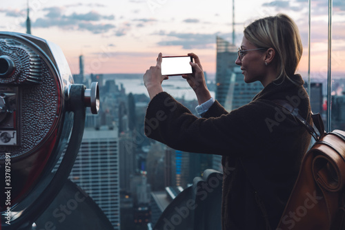 Millennial female traveller using cellphone camera for shooting video content visiting viewpoint observation with coin operated binocular for lookout on New York cityscape,smartphone with blank screen