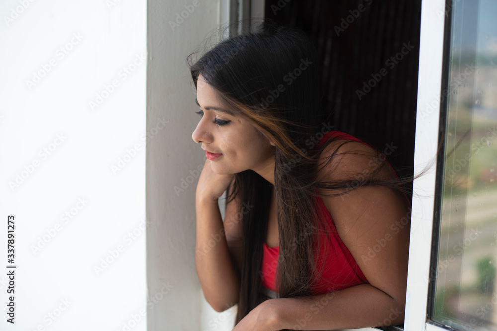Portrait of an young Indian brunette woman in red dress looking outside through a glass window in home quarantine. Indian lifestyle and quarantine. Portrait of an young Indian brunette woman in red dress looking outside through a glass window in home quarantine. Indian lifestyle and quarantine.