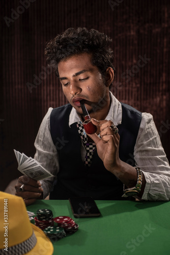 Portrait of young brunette Indian dark skinned man in vintage retro suit playing cards on a casino poker table in front of a textured copy space studio background. lifestyle and fashion.