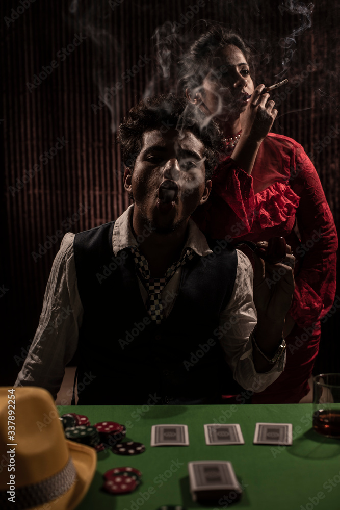 An young and attractive Indian brunette couple in vintage suits smoking in a casino poker table in brown textured copy space background. Gambling and addictive lifestyle. An young and attractive Indian brunette couple in vintage suits smoking in a casino poker table in brown textured copy space background. Gambling and addictive lifestyle.