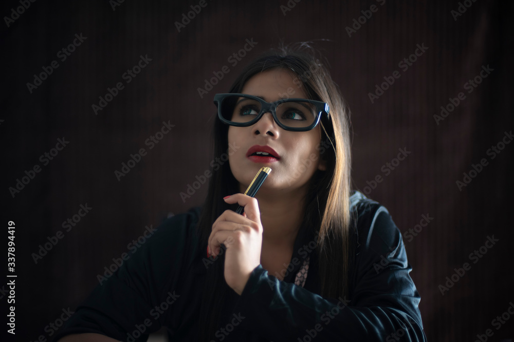 Obraz premium Portrait of an young Indian brunette girl in black corporate suit with a pen in dark copy space studio background. Indian lifestyle and corporate 