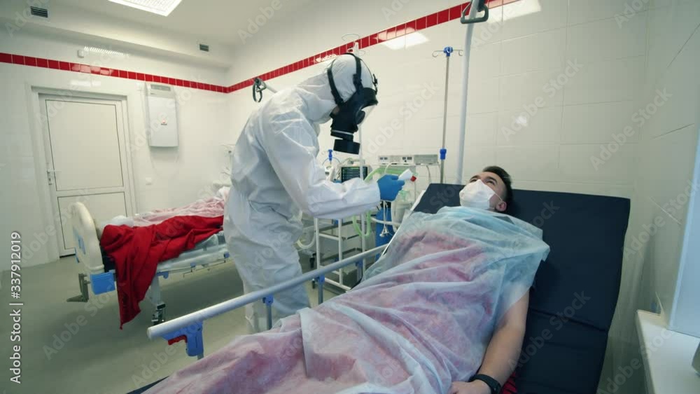 Doctor in a hazmat suit is checking temperature of a lying patient. Covid-19, pandemic, virus, coronavirus disease, world epidemic, coronavirus infection concept. Doctor in a hazmat suit is checking temperature of a lying patient. Covid-19, pandemic, virus, coronavirus disease, world epidemic, coronavirus infection concept.