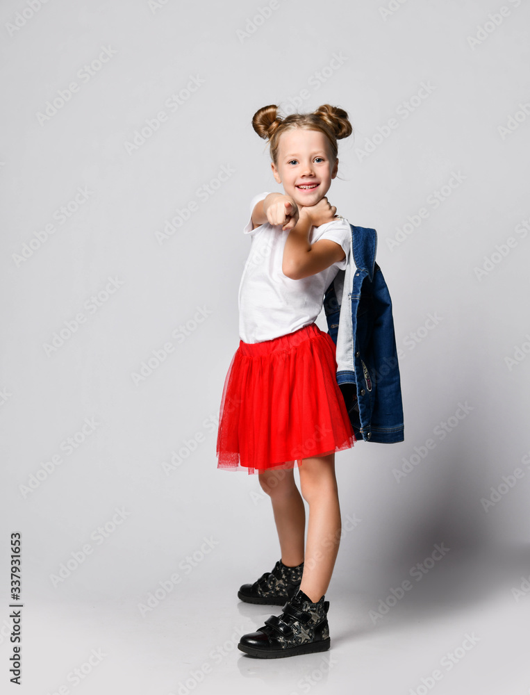 Frolic smiling blonde kid girl with funny buns in red skirt and white t-shirt is pointing finger at us