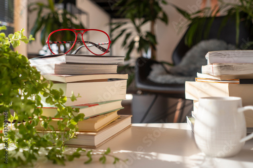 Inside a green house with many plants. A pile of different books on a with table with red glasses and a cup of coffee at sunset