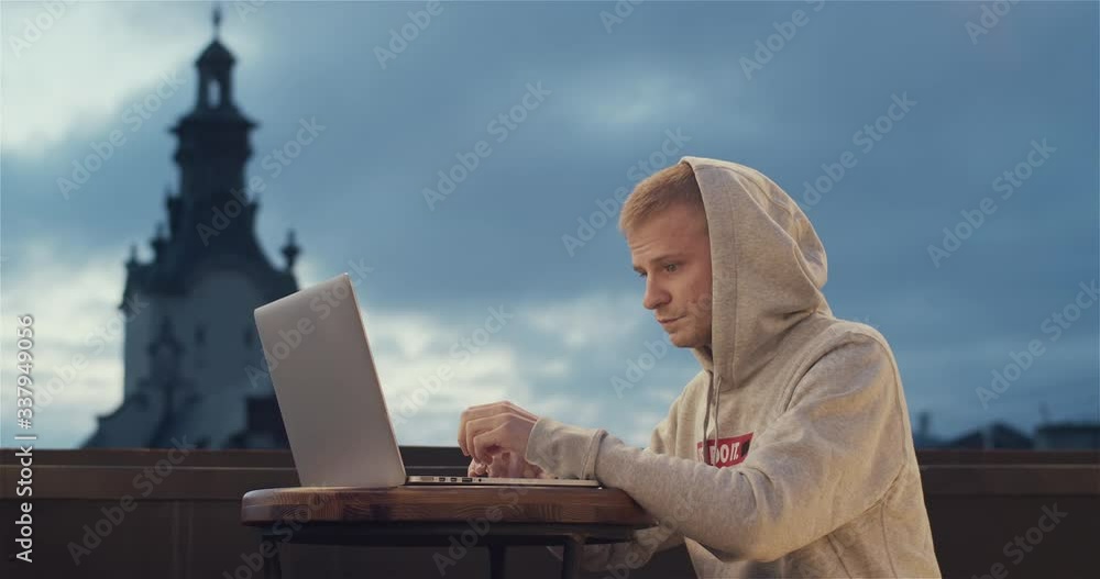 A young man sitting on the roof with a laptop with an old city tower view in the background A young man sitting on the roof with a laptop with an old city tower view in the background
