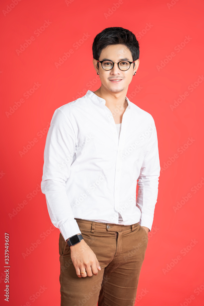 portrait handsome asian businessman with facial hair smiling looking straight, hands in pocket wearing glasses and casual clothing and black earrings, standing and posing with red isolated background portrait handsome asian businessman with facial hair smiling looking straight, hands in pocket wearing glasses and casual clothing and black earrings, standing and posing with red isolated background