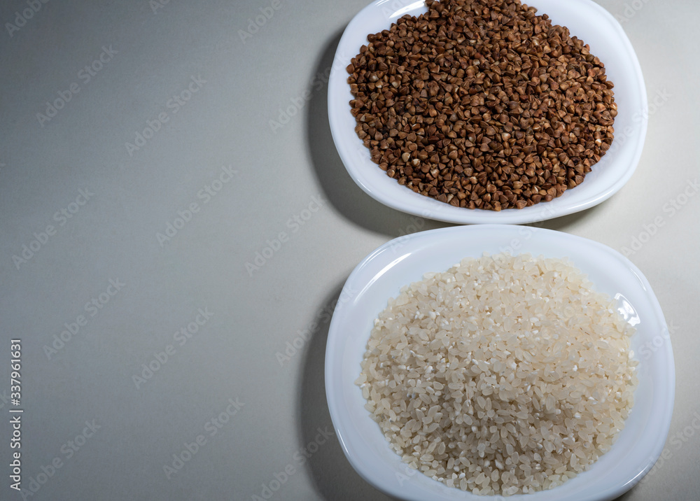 Raw buckwheat grains, uncooked rice on a white plate on a gray background. Ingredient for cooking. View from above. Raw buckwheat grains, uncooked rice on a white plate on a gray background. Ingredient for cooking. View from above.