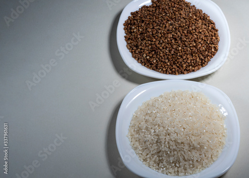 Raw buckwheat grains, uncooked rice on a white plate on a gray background. Ingredient for cooking. View from above.