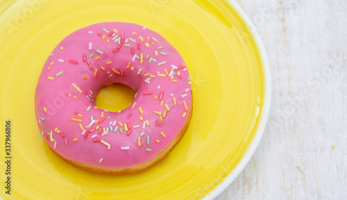 PINK DONUT WITH MULTICOLOR SPRINKLES SERVED IN A YELLOW DISH ON A WHITE WOODEN TABLE