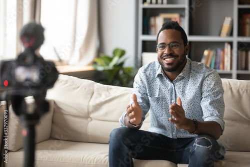 Fototapeta Naklejka Na Ścianę i Meble -  Happy young african american man speaker in eyewear sitting in front of camera, recording educational video webinar training at home. Smiling skilled biracial guy introducing for job interview.