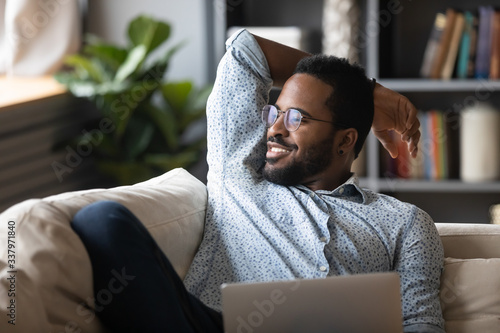 Smiling peaceful handsome african american guy relaxing on comfy sofa with laptop, looking away at window, thinking of opportunities, remembering good life moments, visualizing future at home. photo