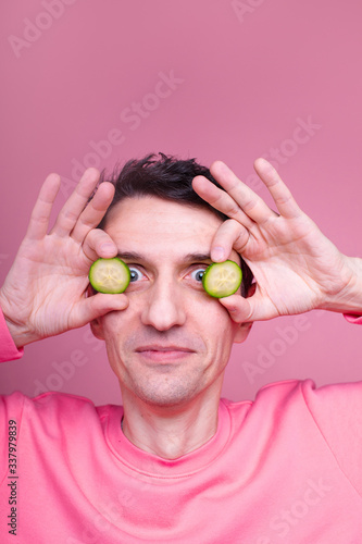 Serious concenrated nice young man hold cucumber slices close to eyes but look straight on camera. Alone in studio. Isolated over pink background.