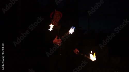 Juggler with a red hat on, performing tricks with 3 burning torches in his hands. Slow motion medium shot.