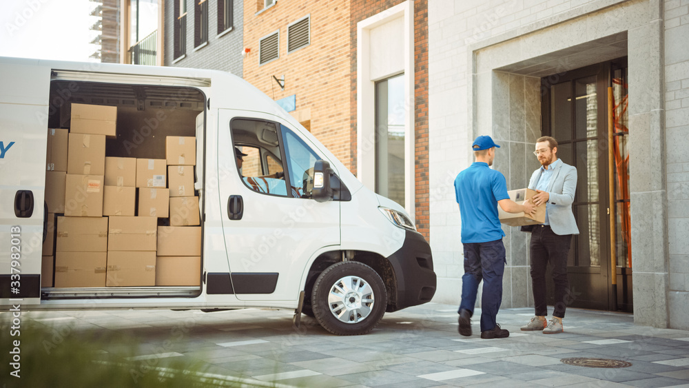 © Gorodenkoff - Delivery Man Gives Postal Package to a Business Customer, Who Signs Electronic Signature POD Device. In Stylish Modern Urban Office Area Courier Delivers Cardboard Box Parcel to a Man