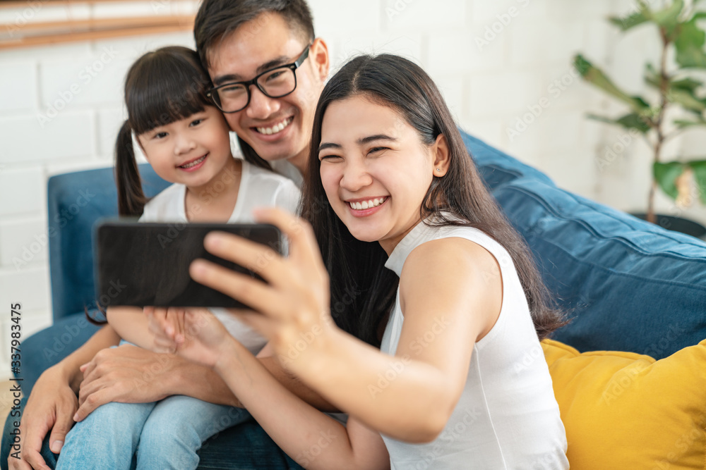 Fototapeta premium Young asian chinese or japanese parents using smartphone tech watch video or enjoy video call in house with kid. Stay healthy, isolate at home on coronavirus outbreak with fun family activity concept.