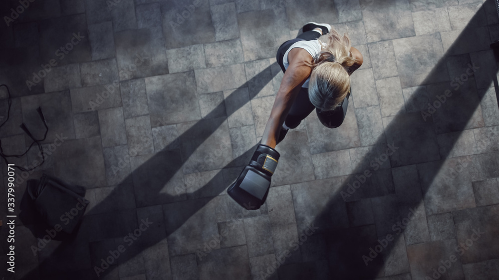 Obraz premium Professional Female Boxer Exercises Air Punches while Wearing Sportswear and Fighting Gloves in the Hardcore Gym. Strong Beautiful Girl Kickboxing and Self Defense Workout. Top Dramatic View