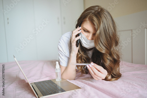 Sick woman in face protection mask lying in bed with laptop and sanitiser holding thermometer at home quarantine isolation. Distance online work from home. Corona virus infection COVID-19 concept.