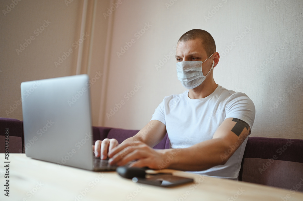 Young european man in face protective medicine mask working on a laptop during coronavirus isolation home quarantine. Covid-19 pandemic Corona virus. Distance online work from home concept.