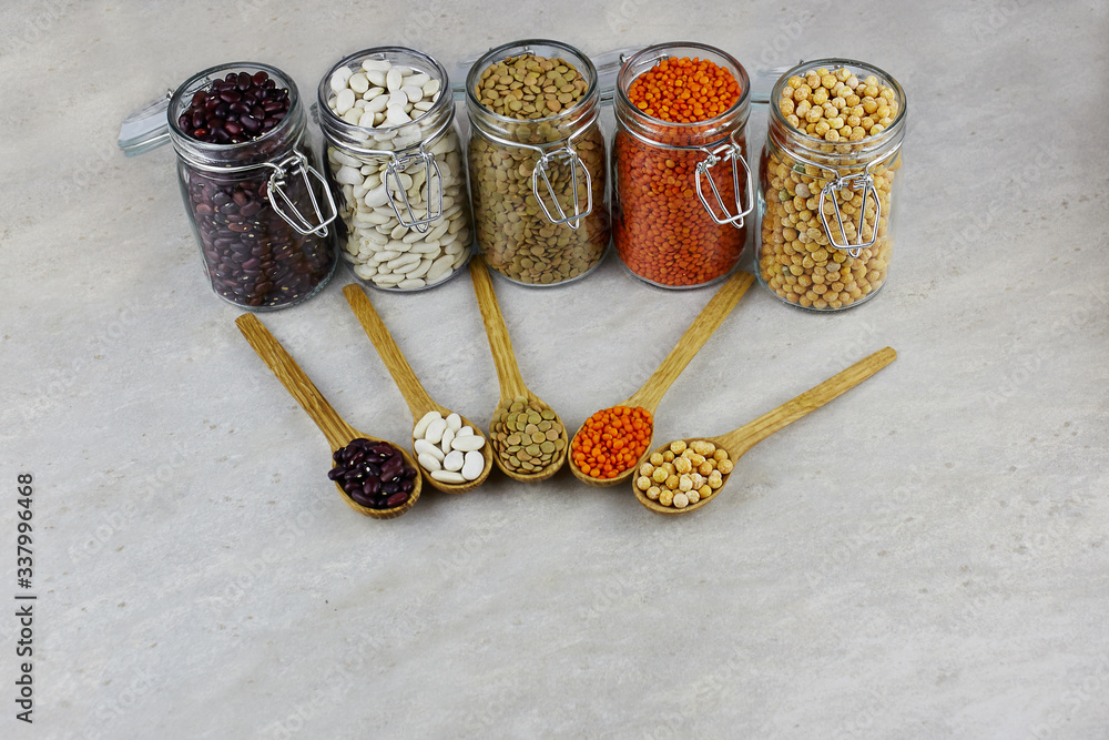 Set of various dried legumes in glass jars and wooden spoons: red lentils, green lentils, peas, red beans, white beans on a gray background Set of various dried legumes in glass jars and wooden spoons: red lentils, green lentils, peas, red beans, white beans on a gray background