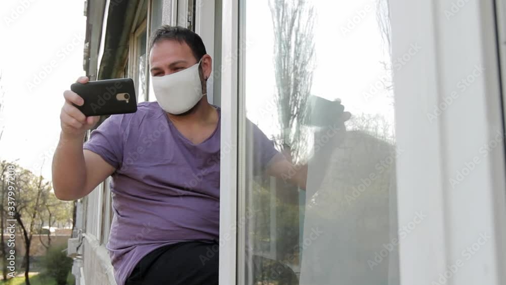 a man with a medical mask on his face sits on a window frame, communicates via video chat in a smartphone, shows a kiss, like, says goodbye, and leaves. camera movement from street to the apartment a man with a medical mask on his face sits on a window frame, communicates via video chat in a smartphone, shows a kiss, like, says goodbye, and leaves. camera movement from street to the apartment