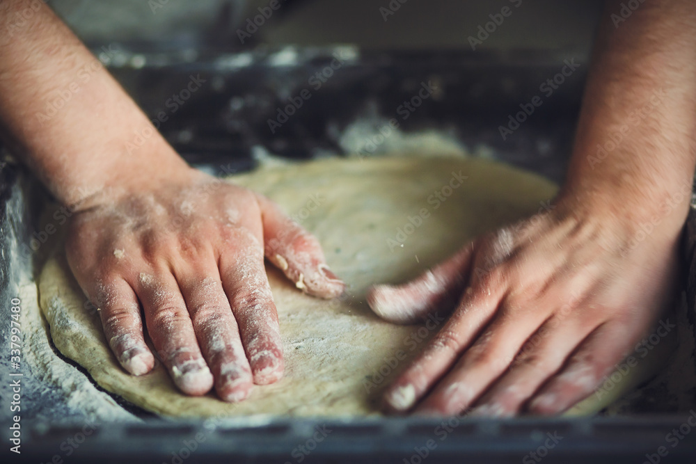 Man's strong hands roll out homemade pizza dough by hand, which lies on a dark baking sheet. Baking at home.