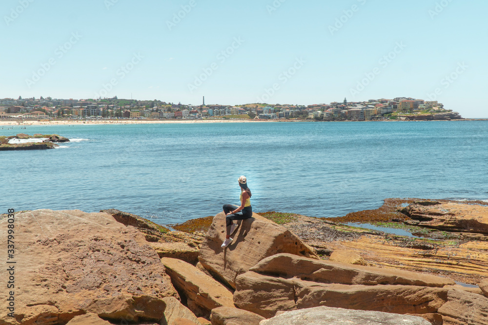 Woman at Bondi Beach. Girl standing on rocks in work out gear looking at view of the ocean, sun, sea and sand scene while on vacation. Holiday, tropical, explore, fitness. Sydney, Australia

