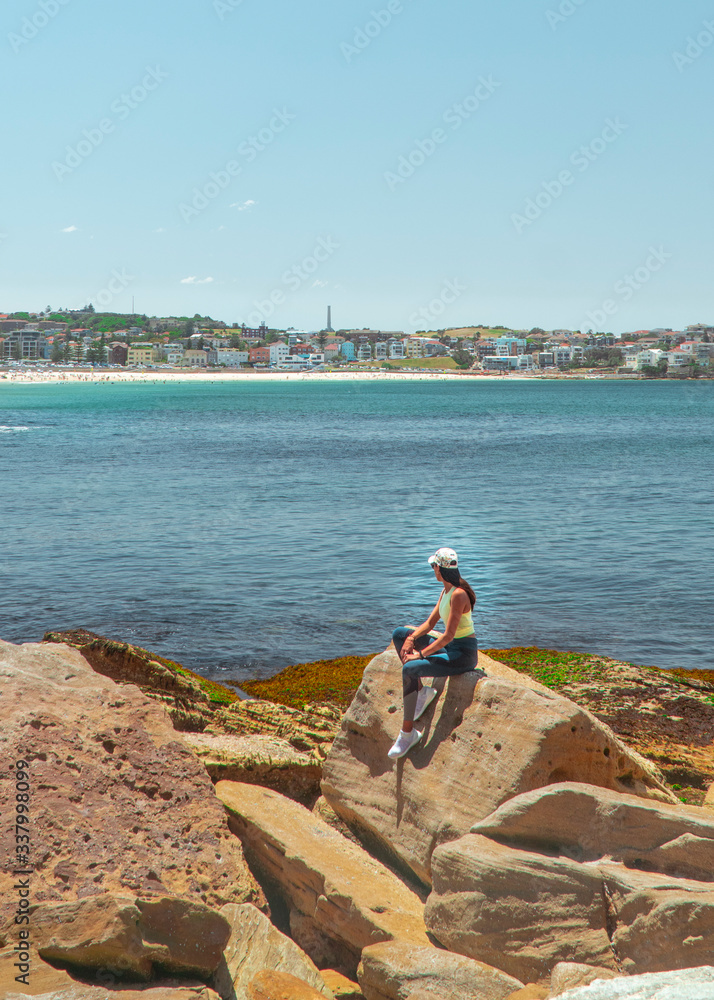 Woman at Bondi Beach. Girl standing on rocks in work out gear looking at view of the ocean, sun, sea and sand scene while on vacation. Holiday, tropical, explore, fitness. Sydney, Australia


