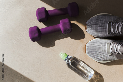 flat lay on a light background: gray sneakers, lilac dumbbells and a bottle of water, hard light, place for text 