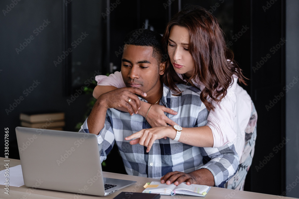 Obraz premium Young asian woman in love embraces her thinking pensive young African American boyfriend working at a computer in a cozy home environment. Pretty wife hugs her successful husband