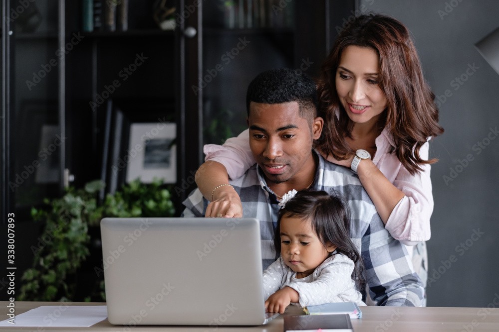 Happy friendly mixed race a family african-american father asian mother and charming daughter are sitting at a table with a laptop and a notebook. Freelance work and happy family concept Happy friendly mixed race a family african-american father asian mother and charming daughter are sitting at a table with a laptop and a notebook. Freelance work and happy family concept