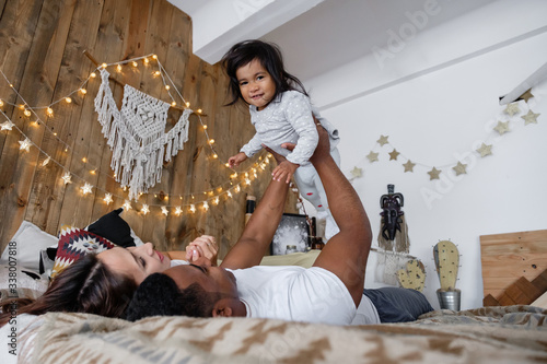 Positive young mixed race family cute young mother African American dad and charming smiling daughter have fun on the bed on Friday evening. Family time concept