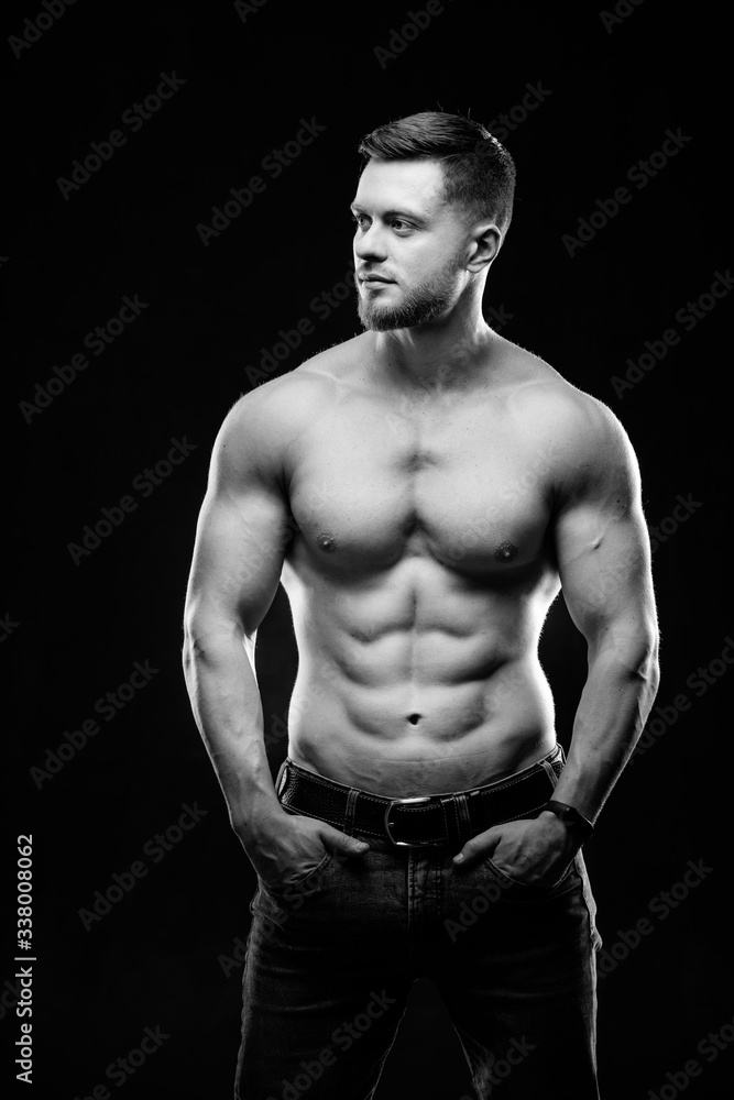 Naklejka premium A portrait of a hot handsome strong guy man without a shirt against dark background. hands in pockets. Studio photo. Male beauty concept. Black and white. Closeup.