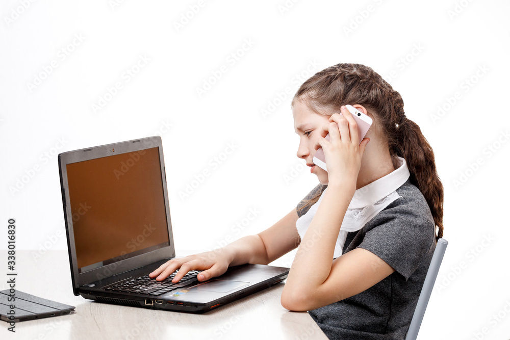 Cute schoolgirl sitting at a laptop and talking on the phone on a white background Cute schoolgirl sitting at a laptop and talking on the phone on a white background